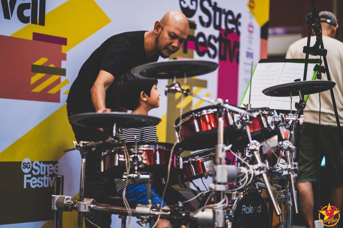 Child playing an electronic drum kit with an adult beside them at a live event.